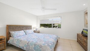 Quarto com uma cama, um ventilador de teto, janela com persianas e uma TV sobre um armário de madeira.
