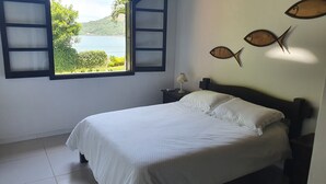 A bedroom with a bed, a nightstand, a lamp, and a window with a view of a body of water and greenery. - Beautiful beachfront house with pool and barbecue area (mangaratiba)