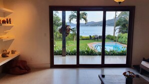 A room with a large sliding glass door leading to a pool area with a view of a lake and mountains. - Beautiful beachfront house with pool and barbecue area (mangaratiba)