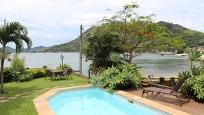 A swimming pool with a view of a lake and hills, surrounded by greenery and a patio area with chairs and a table. - Beautiful beachfront house with pool and barbecue area (mangaratiba)