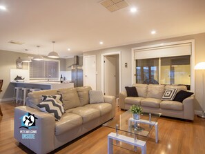 A modern living room with two sofas, a glass coffee table, and a kitchen in the background. - Stylish 3BR Lake- front House + Kayaks (Encounter Bay)