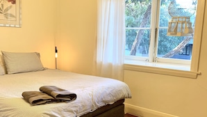 A bedroom with a bed, a bedside lamp, a window with curtains, and a view of trees and a birdhouse outside. - Free parking & 3 min walk to beach! (Bondi Beach)