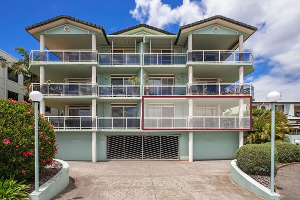 Un edificio de varios pisos con balcones, entrada de garaje y zonas verdes circundantes.