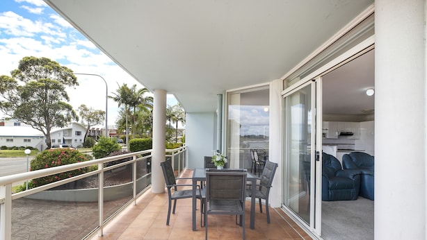A balcony with a table and chairs, a glass door leading to a living area, and a view of the street and greenery.