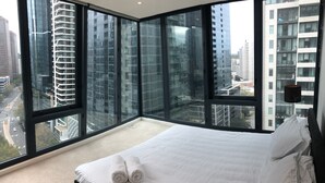 Une chambre à coucher avec un grand lit, du linge de lit blanc et une vue sur les grands immeubles, grâce à de larges fenêtres du sol au plafond.