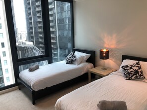 Ein Hotelzimmer mit zwei Betten, einem Nachttisch mit Lampe und einem großen Fenster mit Blick auf die Stadt.