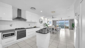 A modern kitchen with white cabinets, a black countertop, and stainless steel appliances. There is a dining area with a table and chairs, and a large window offering a view of the outdoors. - Lagoon 206 Hamilton Island by HamoRent (Hamilton Island)