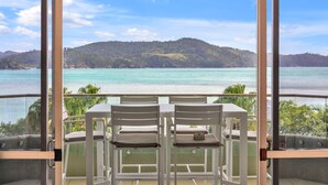 A balcony with a table and chairs overlooking a scenic view of a lake and forested hills. - Lagoon 206 Hamilton Island by HamoRent (Hamilton Island)