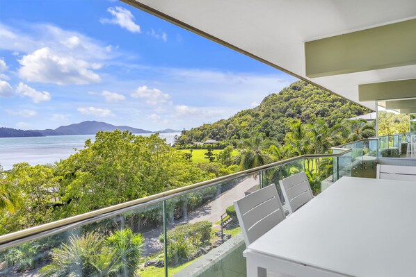 A balcony with a glass railing overlooking a lush green landscape and a body of water. - Lagoon 206 Hamilton Island by HamoRent (Hamilton Island)