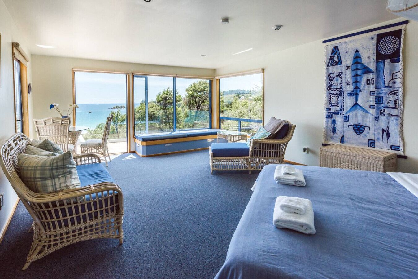 A bedroom with a bed, two wicker chairs, a bench, and a view of the sea.