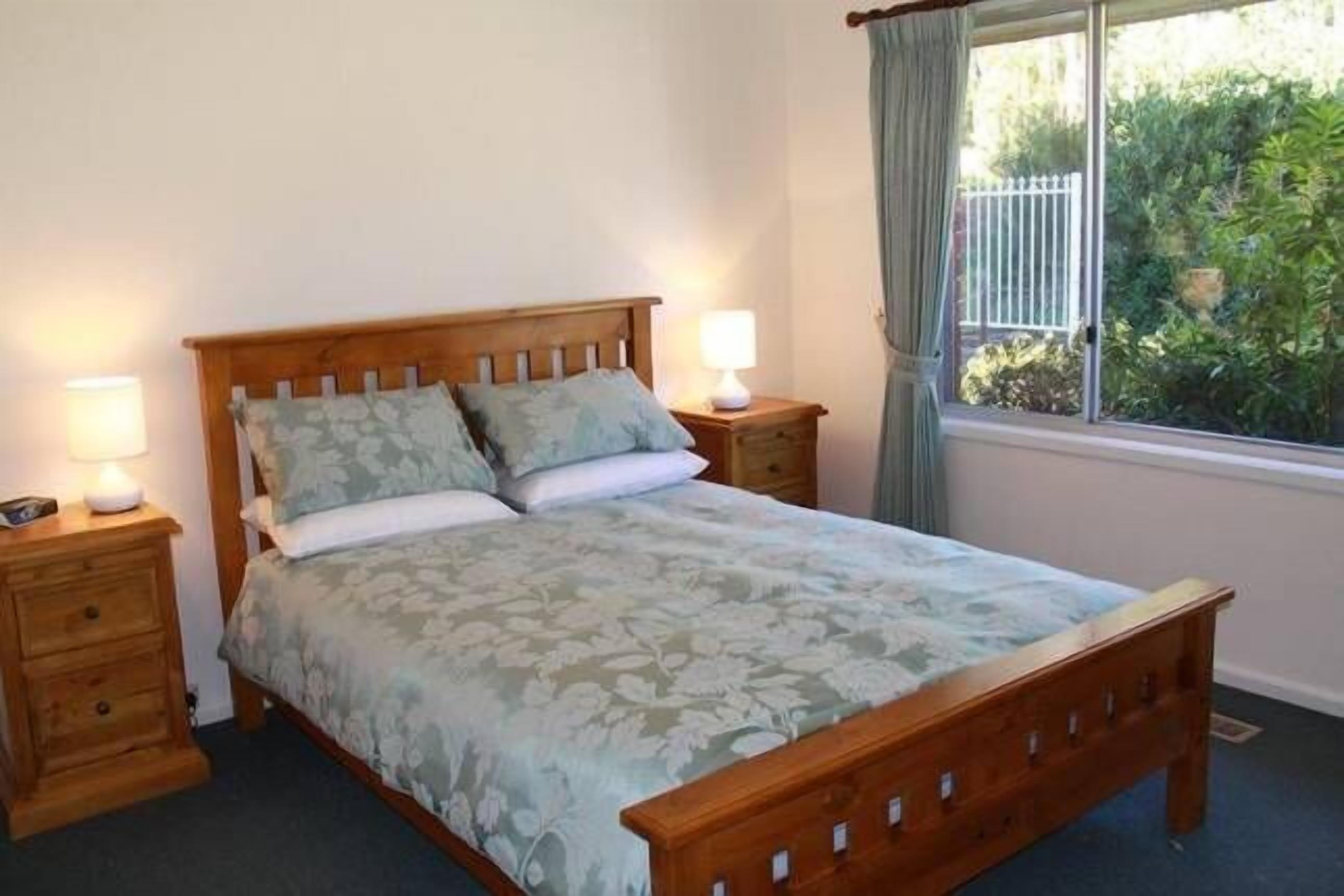 Un dormitorio con cama de madera, dos mesitas de noche, una ventana con cortinas y vistas a la vegetación.