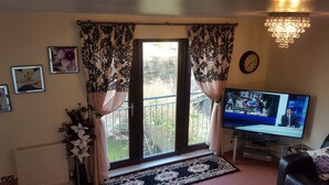 Ein Wohnzimmer mit Fernseher, der eine Nachrichtensendung zeigt, einem Fenster mit Vorhängen, einer Heizung und einer Kristallleuchtre.