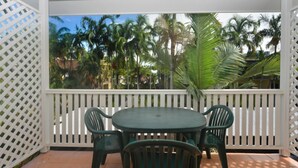 Un porche cubierto con una mesa redonda y cuatro sillas, rodeado por una cerca de celosía blanca, con vistas a un jardín tropical con palmeras.
