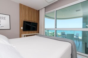 A hotel room with a large bed, a flat-screen TV mounted on a wooden panel, and a balcony with a glass railing offering a view of the city and ocean.