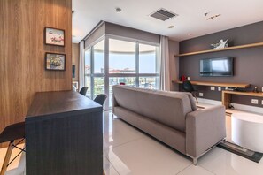 A modern living room with a sofa, a coffee table, a TV, and a large window offering a city view.