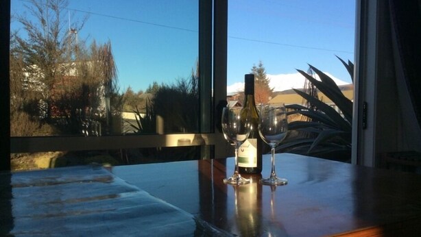 Een tafel met een fles wijn en twee glazen, een raam met uitzicht op bomen en een gebouw, en een houten tafelrand.