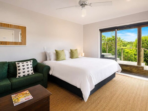A bedroom with a bed, a green sofa, a wooden coffee table, and a large window with a view of greenery.