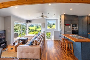 A modern kitchen with a wooden countertop and built-in appliances, a dining area with a sofa and a television, and large windows offering a view of the outdoors.