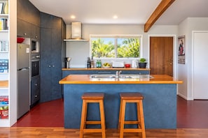A modern kitchen with a central island, wooden stools, a built-in oven, and a window with a view of greenery.