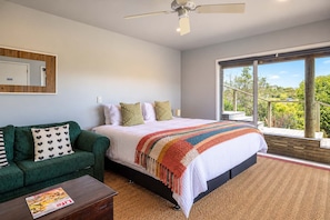 A bedroom with a bed, a green sofa, a wooden coffee table, and a large window with a view of greenery.