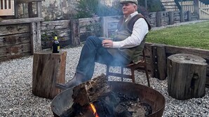 A man sitting by a fire pit in an outdoor setting with a rustic stone building in the background. - Christmas 2026 Availability (Bundanoon)