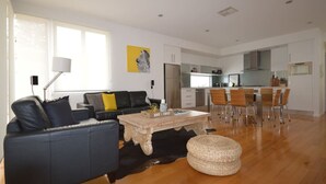 A modern living room with a sofa, coffee table, and ottoman. There is a dining area with chairs and a table. The kitchen is visible in the background with cabinets and appliances. - The Holdfast Townhouse Sussex St Glenelg (Glenelg)