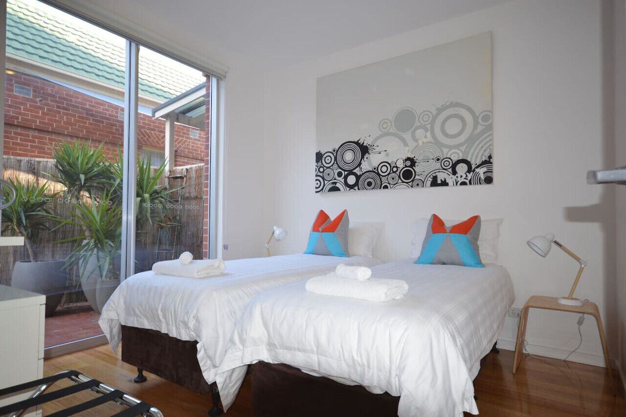 A modern hotel room with two beds, a large window, and a view of a brick building. - The Holdfast Townhouse Sussex St Glenelg (Glenelg)