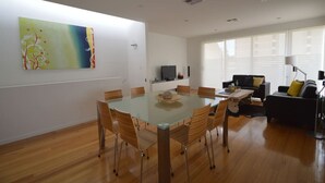 A modern living room with a glass dining table, wooden chairs, a black sofa, and a large abstract painting on the wall. - The Holdfast Townhouse Sussex St Glenelg (Glenelg)