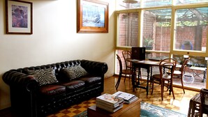 A living room with a leather sofa, a coffee table with magazines, a dining table with chairs, and a large window with blinds. - Stanley Cottage 2 bedroom (North Adelaide)