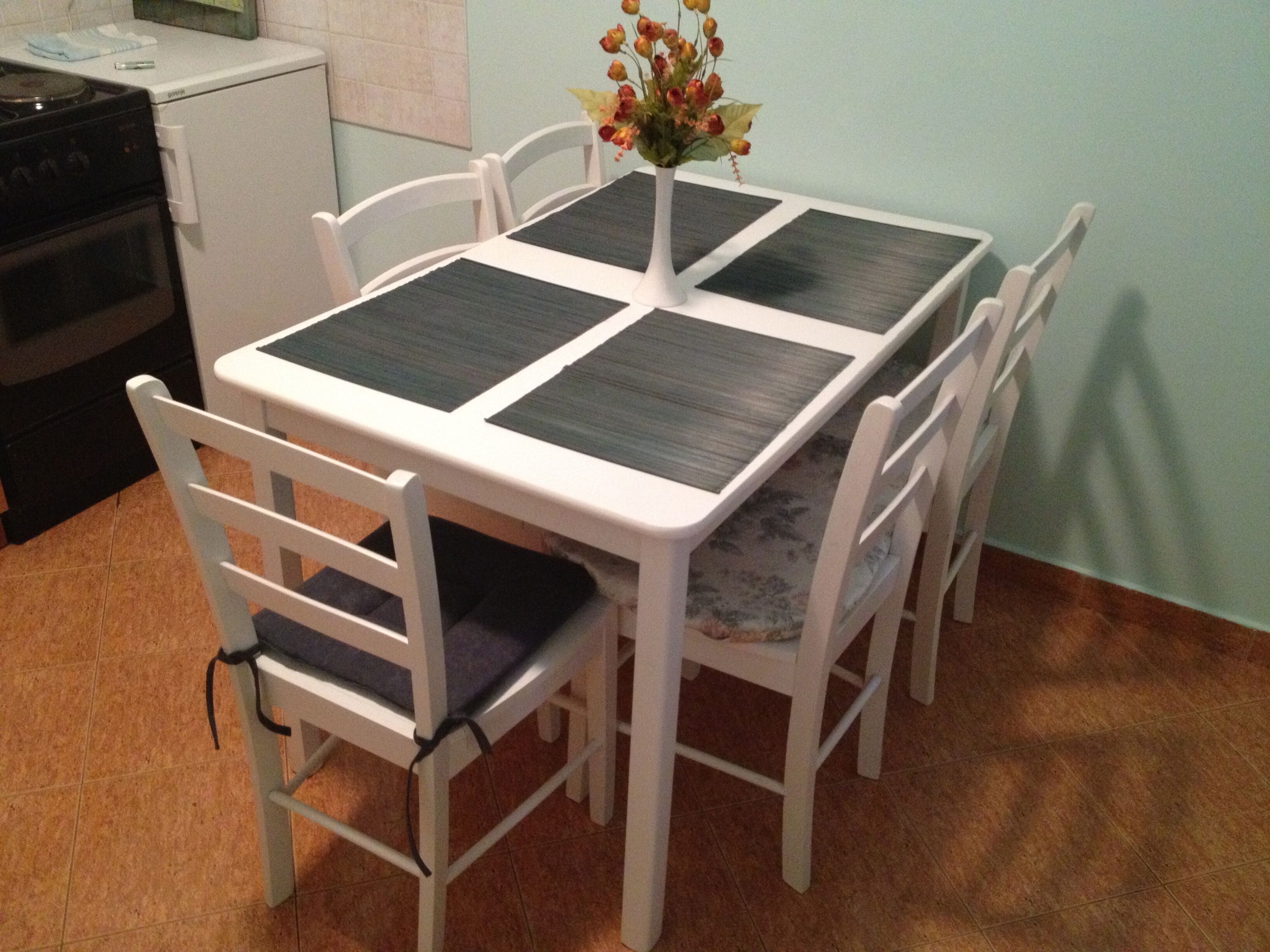 A dining table with four chairs, a vase with flowers, and a kitchen area with a stove and refrigerator.