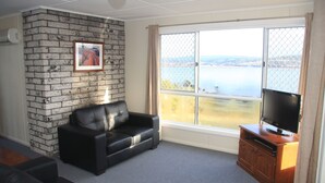 A living room with a black leather sofa, a TV on a wooden cabinet, a large window with a view of a body of water, and a brick wall. - API Leisure & Lifestyle St Helens (St Helens)