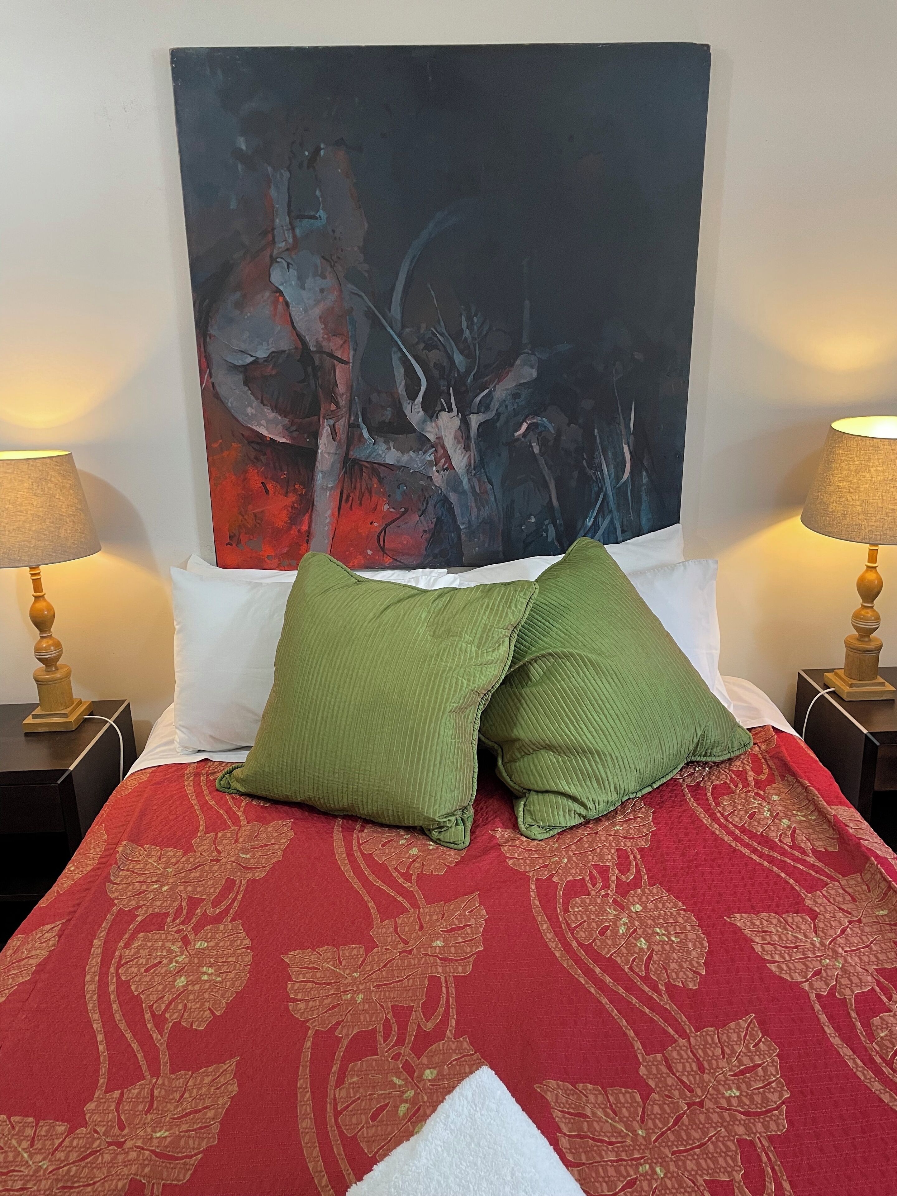 A bed with a red floral bedspread, two green pillows, and a large abstract painting on the wall behind.