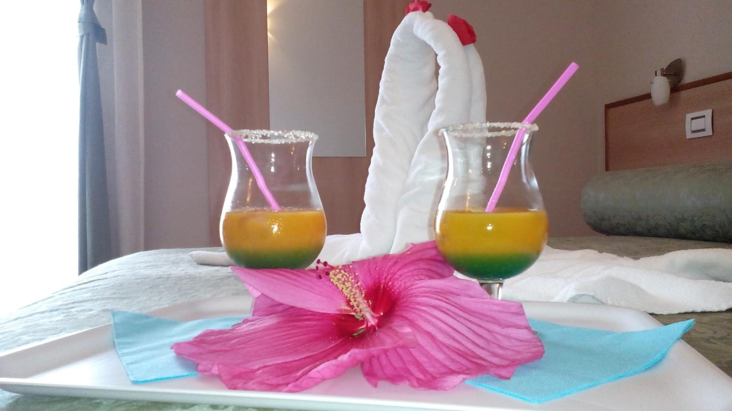 Two drinks in glasses with pink straws on a tray, a large pink flower, and a white towel on a bed.