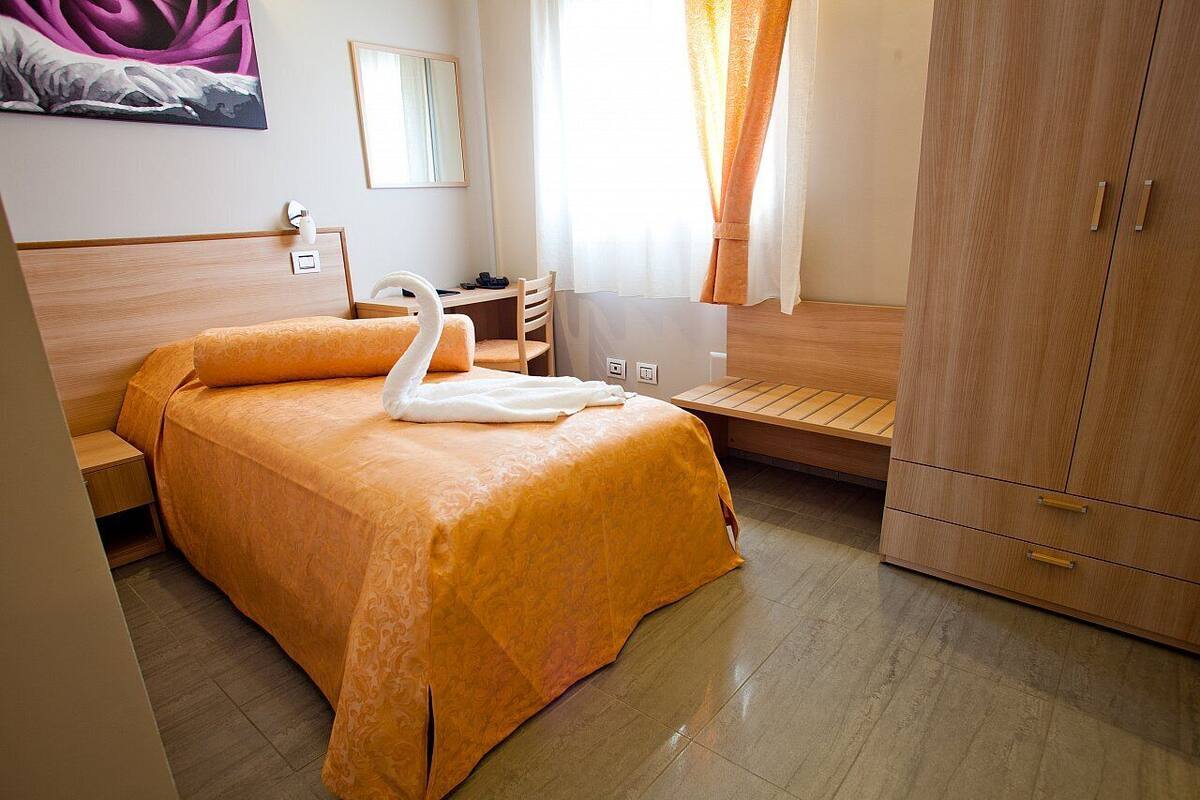 A hotel room with a bed, a desk, a chair, a wardrobe, and a painting on the wall.