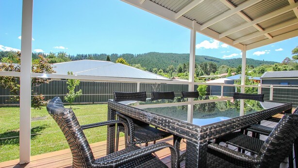 A covered patio with a glass-top table and wicker chairs, overlooking a green landscape and distant hills. - BLUE HILLS RISE: Bright (Bright)