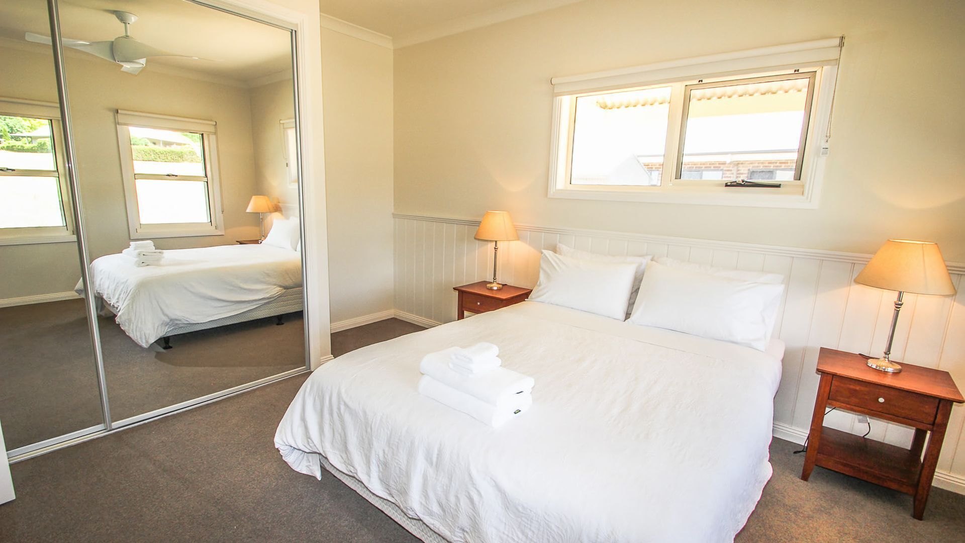 Una cama bien hecha con ropa blanca y toallas, dos mesitas de noche con lámparas, una ventana con cortinas y un espejo que refleja la habitación.