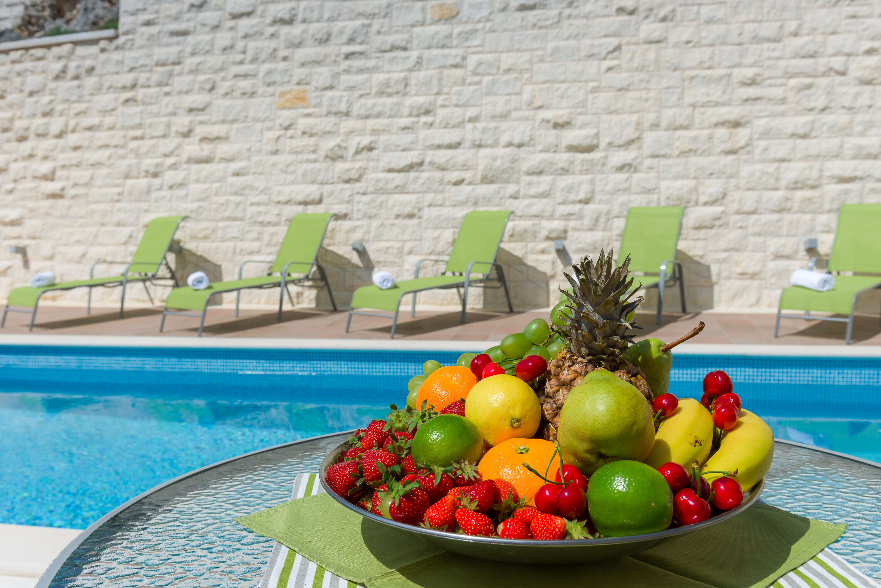 A fruit bowl on a table by a pool with green chairs in the background.