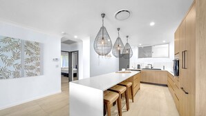 A modern kitchen with a white island, wooden bar stools, pendant lights, and wooden cabinetry. - The Elite F208 Hamilton Island by HamoRent (Hamilton Island)
