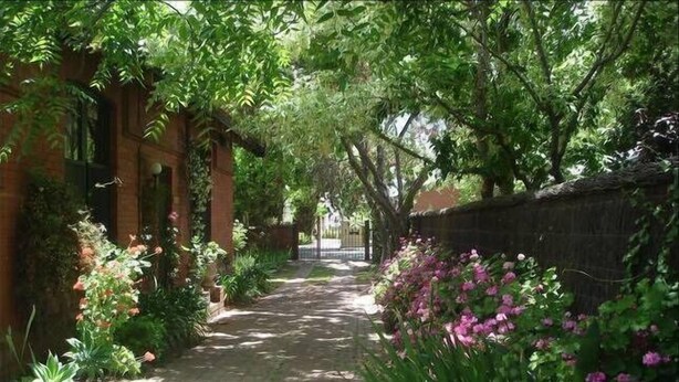 Un sentier bordé d’une végétation luxuriante et de plantes en fleurs, qui mène à un bâtiment aux murs de briques et à l’entrée abritée.