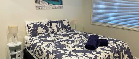 A bedroom with a large bed, a nightstand with a lamp, a wall-mounted artwork, and a window with blinds.