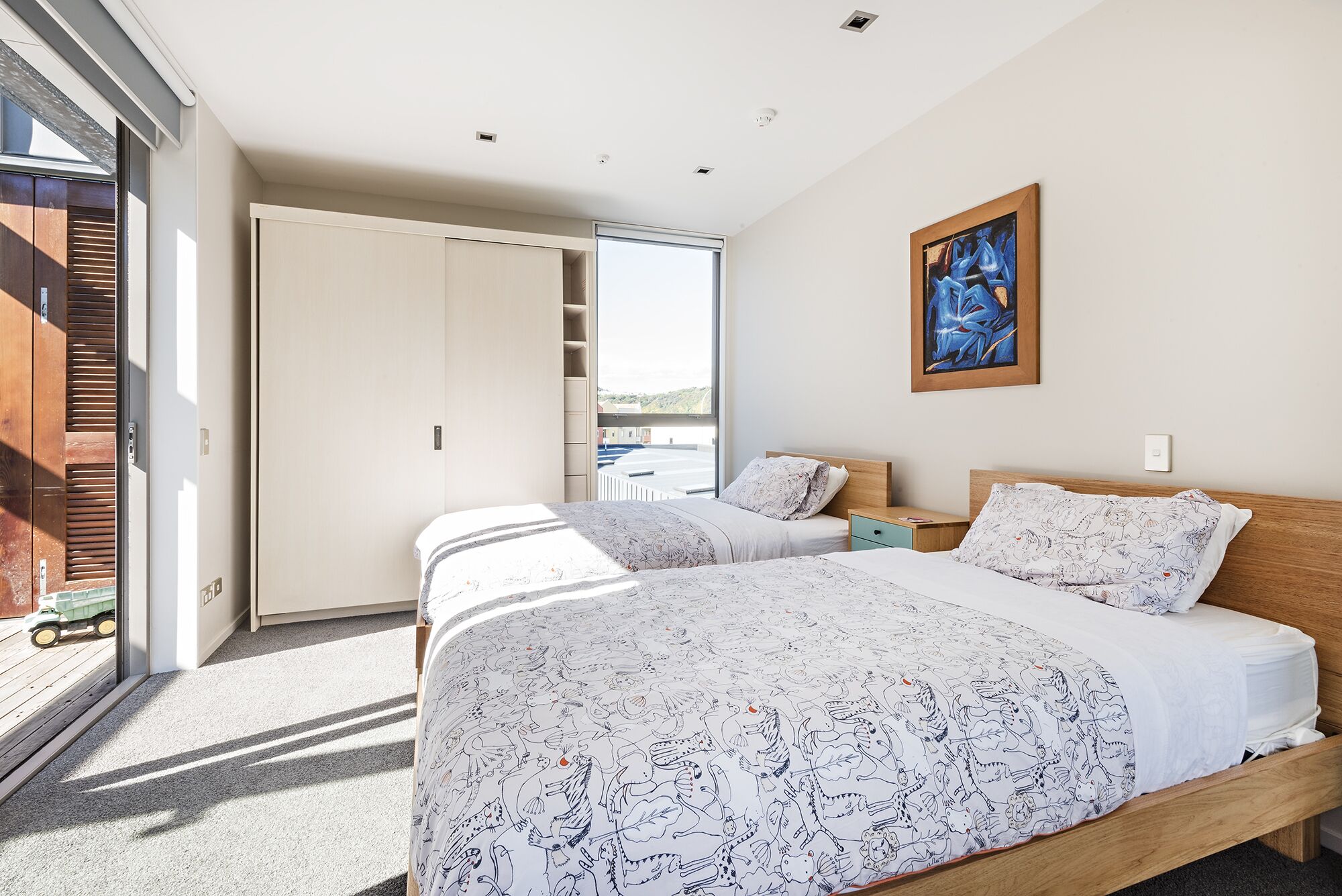 A bedroom with two beds, a large window, and a painting on the wall.