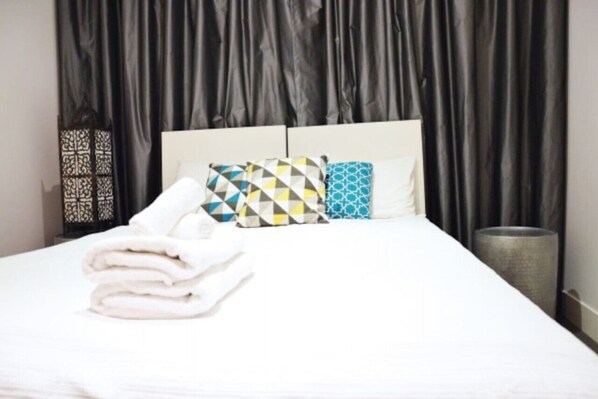A neatly made bed with white linens and two patterned pillows, a black curtain backdrop, and a decorative wall piece to the left.