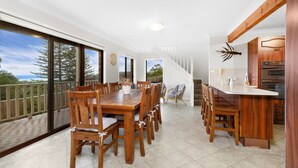 A spacious dining area with a large wooden table and chairs, a kitchen with wooden cabinets, and a balcony with a view of the ocean. - Shirl's By The Sea - beach at your doorstep (Gerroa)
