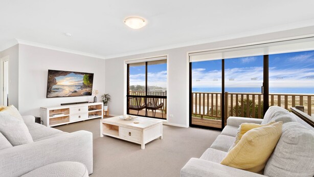 A spacious living room with a large TV, a white sofa, a coffee table, and a balcony with a view of the beach. - Shirl's By The Sea - beach at your doorstep (Gerroa)