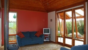 Ein Zimmer mit einem blauen Sofa, einem Fernseher auf einem Schrank, einem großen Fenster mit Blick auf Grünflächen und einer Holzzieldecke.