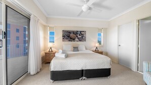 A bedroom with a large bed, a ceiling fan, two bedside tables with lamps, a window with a view, and a door.