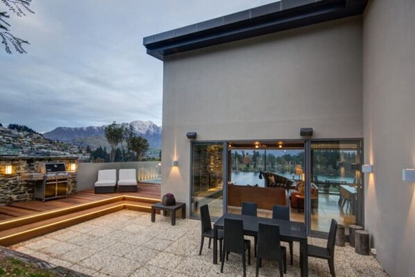 A modern outdoor patio with a stone barbecue, seating area, and glass doors leading to a pool.