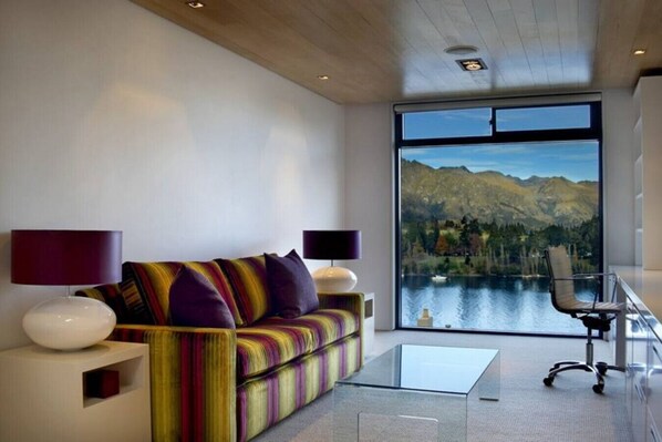 A modern living room with a sofa, two lamps, a glass coffee table, and a view of a lake and mountains. - Lakeview by Sotheby's Luxury Rental Homes (Queenstown)