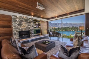 A modern living room with a stone fireplace, leather sofa, and a large window offering a mountain lake view. - Lakeview by Sotheby's Luxury Rental Homes (Queenstown)