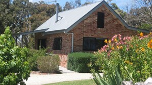 A brick house with a metal roof, surrounded by greenery and flowering plants. - Prom Coast Holiday Lodge - Cottage 1 (Waratah North)